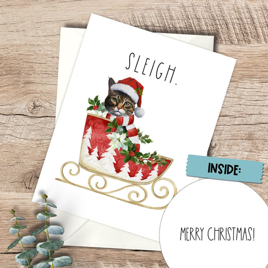 Christmas card with a cat in a Santa hat and sleigh on a wooden surface.